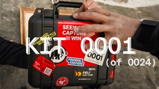 A person holding a black Peli 1200 case with red and yellow stickers, labeled "KIT 0001 (of 0024)" against a textured wall