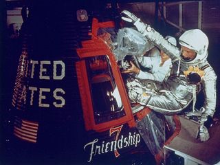 A light-skinned man in a silver spacesuit grips above the window of his small, black, Friendship 7 space capsule. Red tape frames the windows, and his legs are tucked into an opening where a hatch would be as he hangs off the side. A technician dressed in white handles a component above Glenn's legs. The last half of the words "United States" are seen printed on the capsule to the left of Glenn, underneath is an american flag. "Friendship 7" is painted beneath the window.