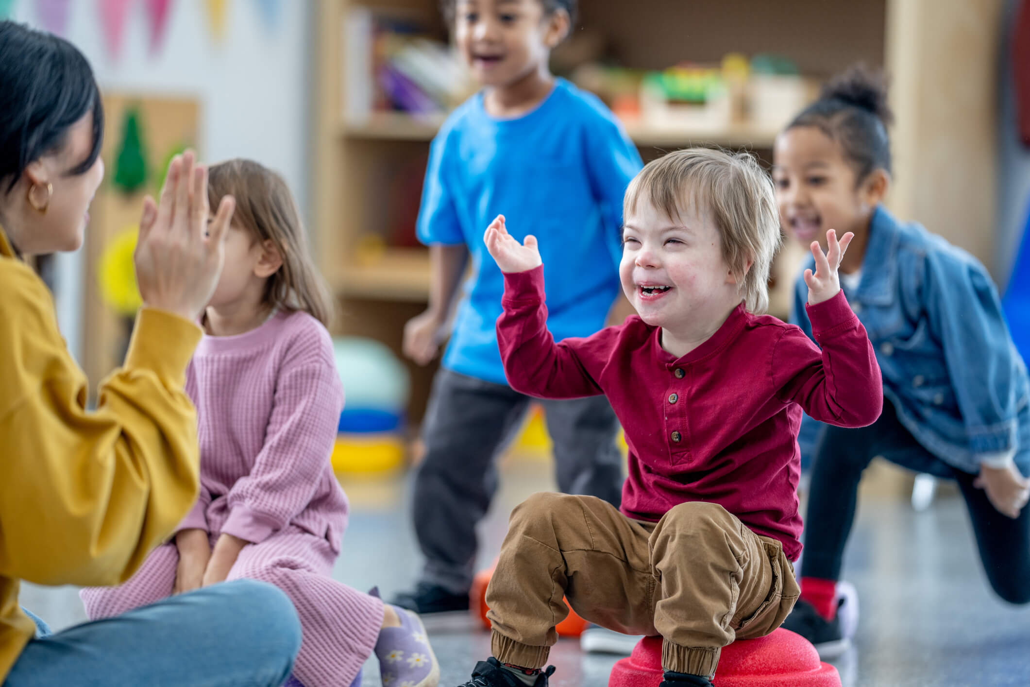 A small group of preschool children sit on the floor in front f their teacher as she leads them in a song. The children are singing along and doing the actions as they follow the teachers lead.