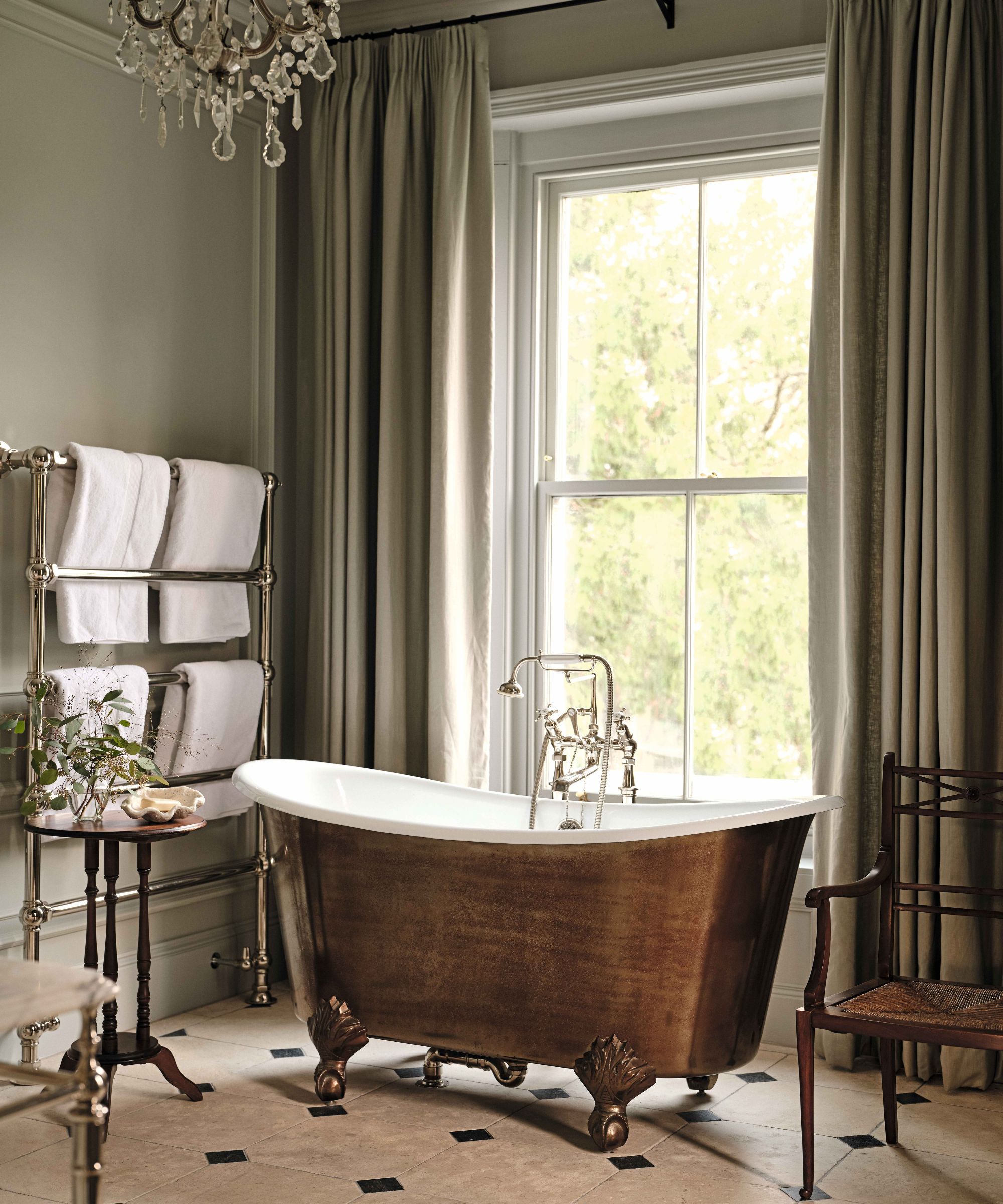 a moody green bathroom with large floor to ceiling georgian windows, a copper freestanding bathtub, chandelier, stone floors and antique furniture