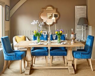 Blue upholstered dining chairs in French-country dining scheme, with solid wood dining table, shapely mirror on wall, earthy neutral walls, and fresh yellow flowers in vases.
