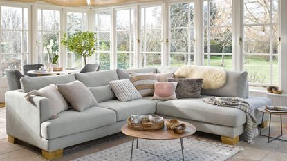 large conservatory with pale grey L-shaped sofa, small wood and metal coffee table, dining table and chairs in background, ceiling blinds, cushions, sheepskin, throw, rug, tiled floor, view of garden