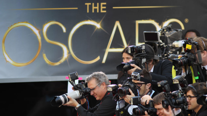 Photographers snap stars on the red carpet in front of a large backdrop that says 'The Oscars'