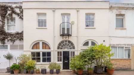 the exterior of the house, which has a white brick front with three square windows across the top floor, and two wide rounded windows on the ground floor either side of a black painted door. There are potted plants dotted around the front