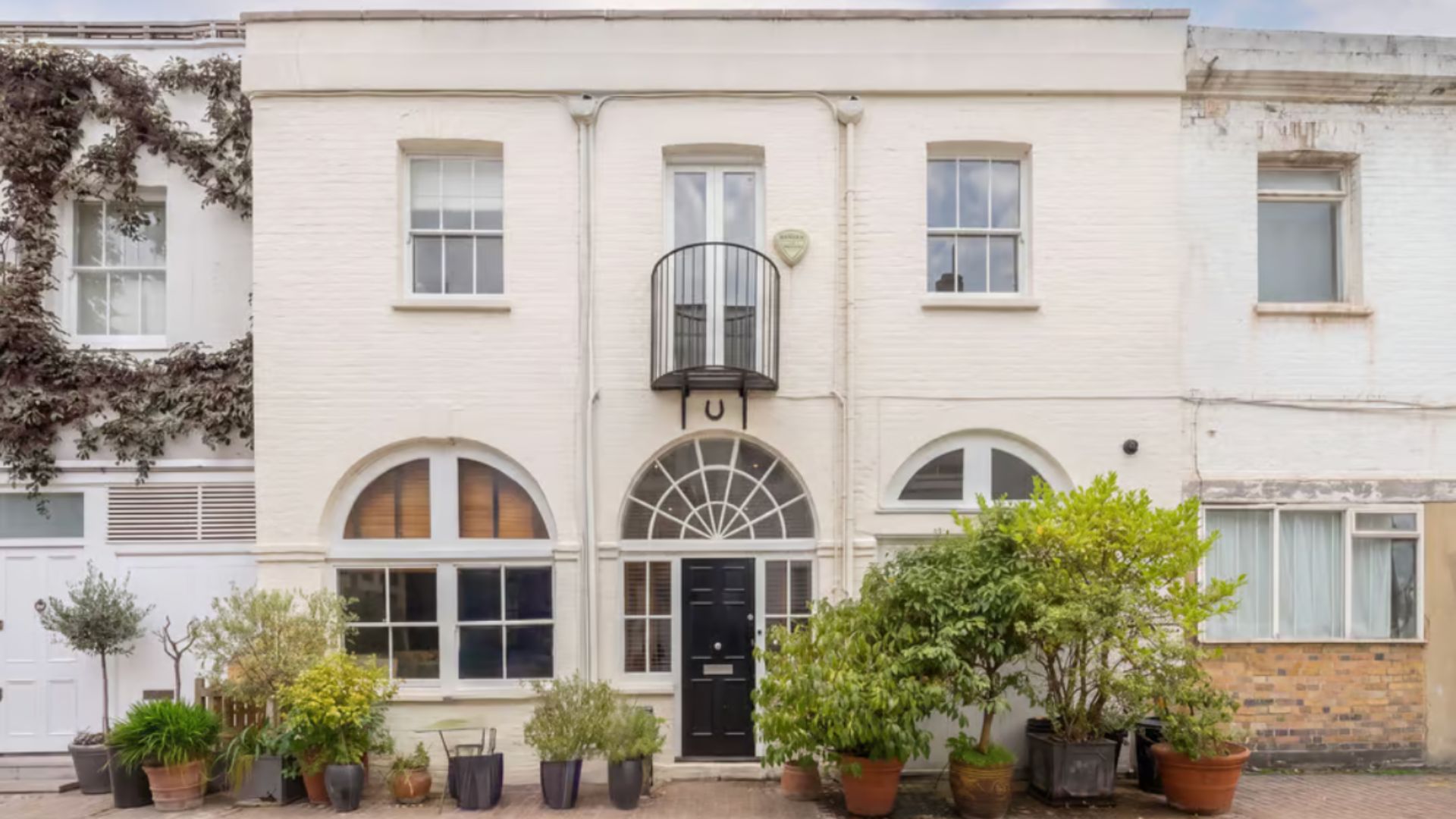 the exterior of the house, which has a white brick front with three square windows across the top floor, and two wide rounded windows on the ground floor either side of a black painted door. There are potted plants dotted around the front 