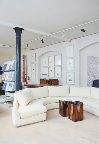 The modern interiors of a sun-lit shop feature framed artworks, a semicircular sofa, wooden coffee tables, and a zig-zag book shelf.