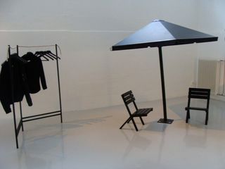 A room featuring white walls and floor. In the left of the room is a black hanging rail with black hangers and black jackets hanging off it. On the right of the room is a black parasol with 2 black chairs