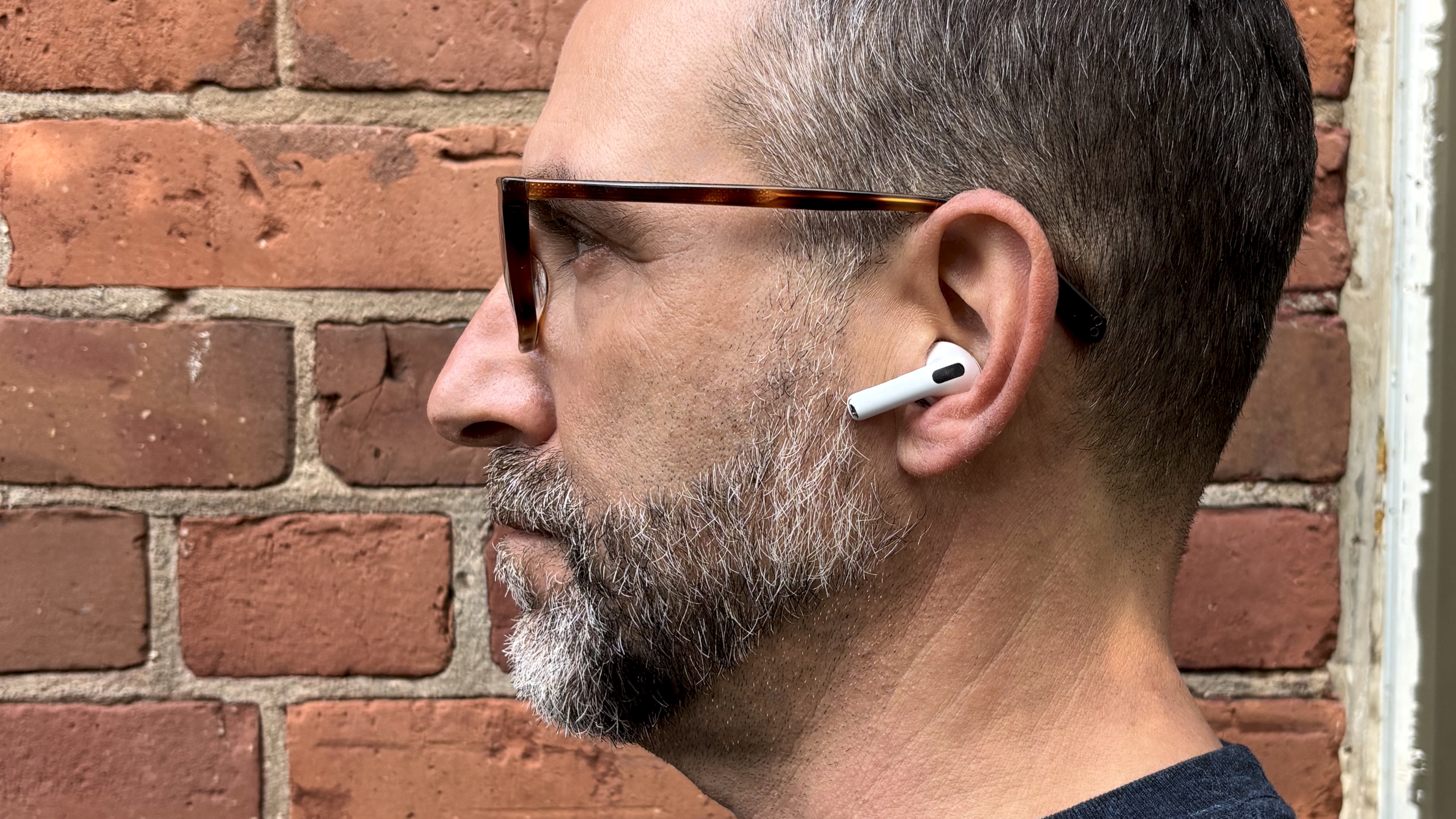 Apple AirPods Pro 3 either worn by a man standing in front of a red-brick wall, or on a table with gray twine cover 