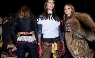 Four female models wearing looks from the Dsquared2 collection. One model is wearing a dark coloured piece with multicoloured detail and fringe trims. Next to her is a model wearing a short black jacket, a multicoloured piece with white lace hem and blue and red trousers. The third model is wearing a white top with a black bow and lace detail with denim sleeves showing through and brown trousers with a brown and red layer on top and yellow tassels. The fourth model is wearing a dark coloured piece and she is holding a brown and green camouflage style jacket with brown fur