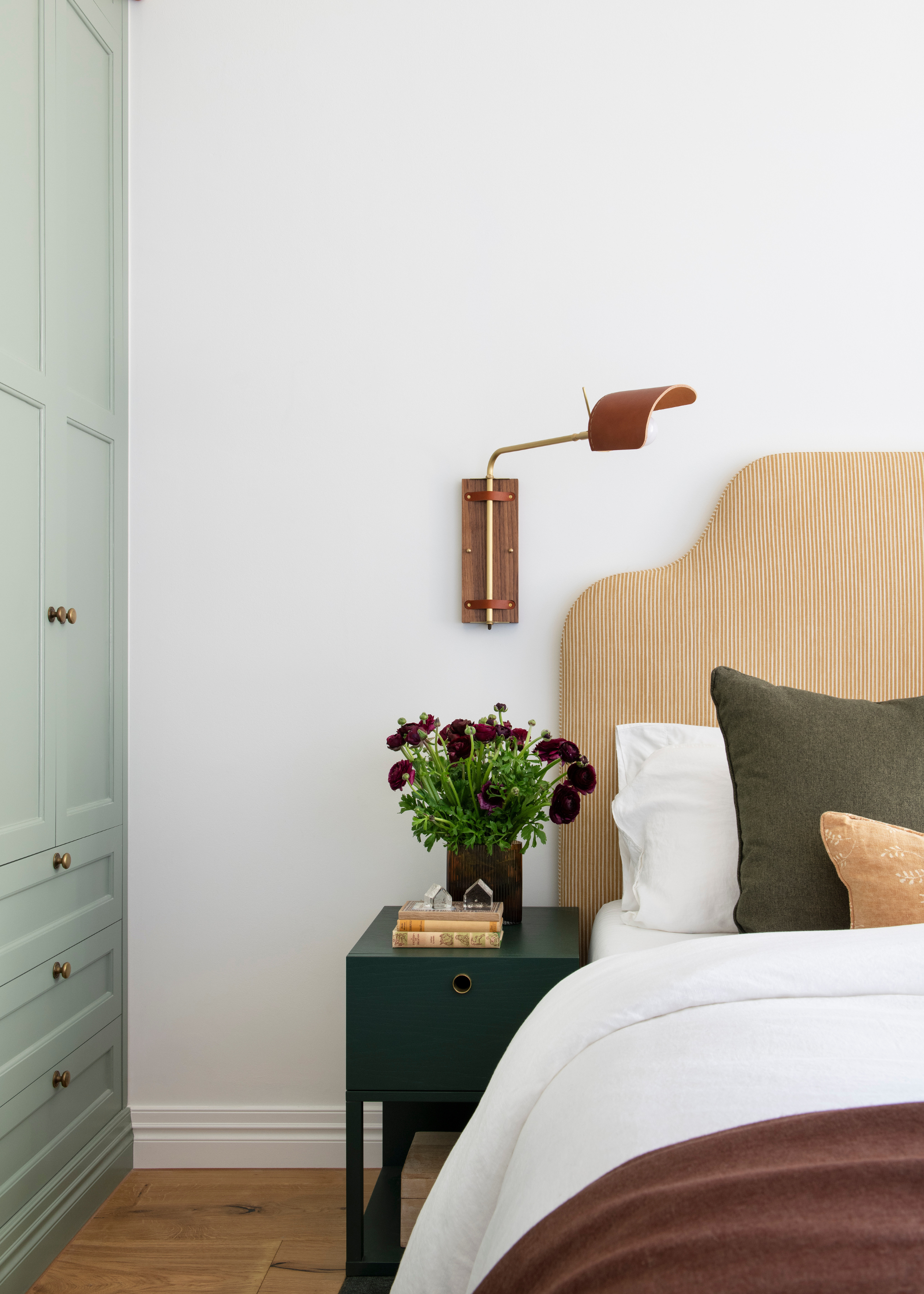A white bedroom with a wood wall sconce, a striped headboard, a black side table with a vase of flowers, and green cupboards