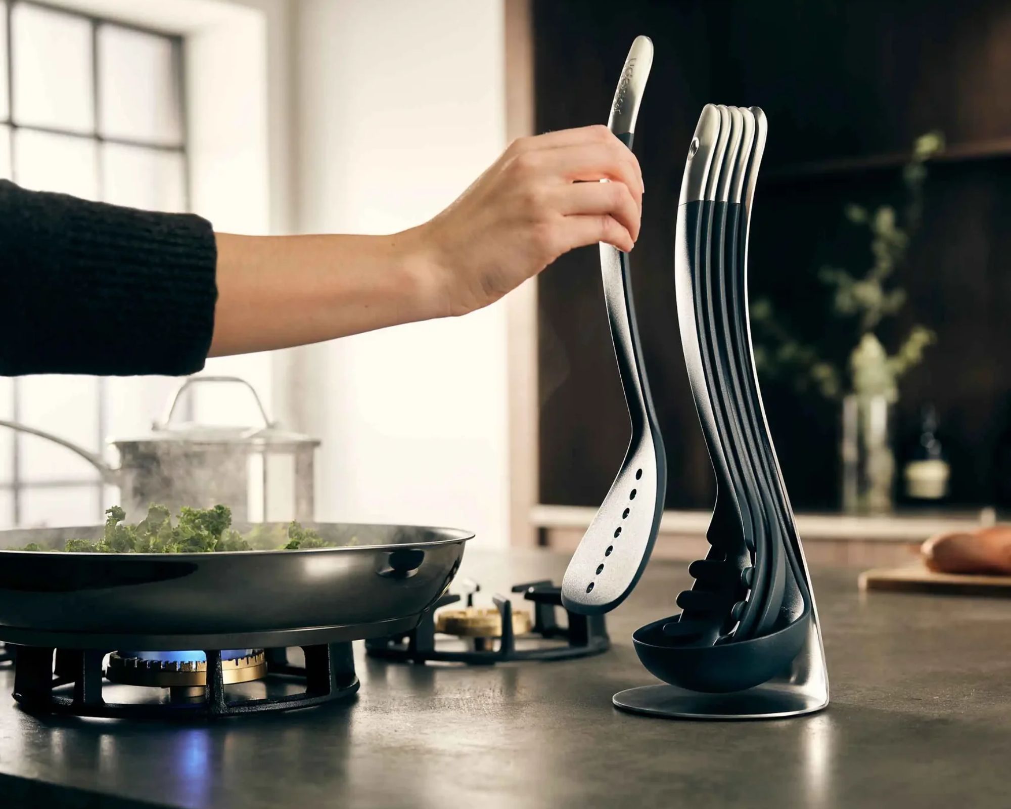 A hand reaching for the JJ Nesting utensil storage stand while cooking green vegetables in a silver frying pan over a gas cooker in a neutral kitchen.