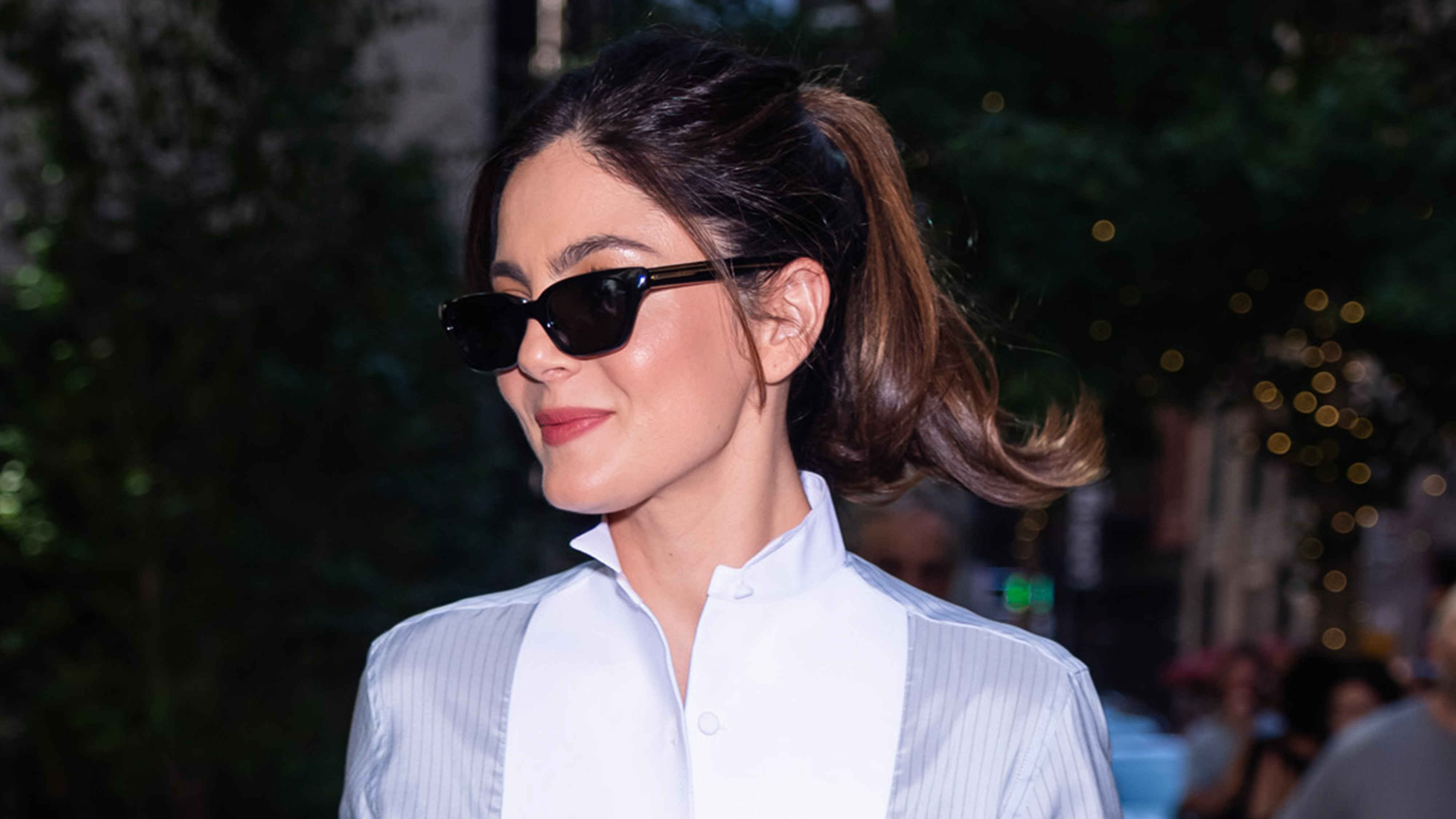 monica barbaro wears a tuxedo shirt and sunglasses.