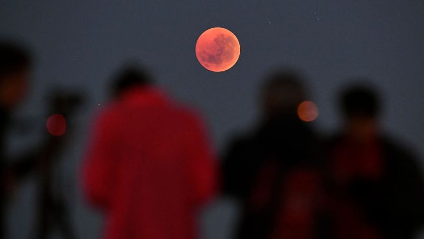 people stand in foreground and look at blood red moon in the sky as the total lunar eclipse takes hold. 