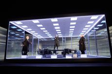 The men perform in a mocked up office enclosed in a large glass cube.