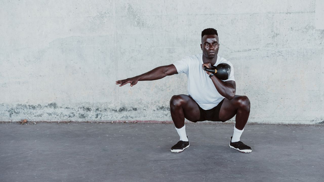 Sportsman with arms outstretched holding kettle bell while crouching against wall 