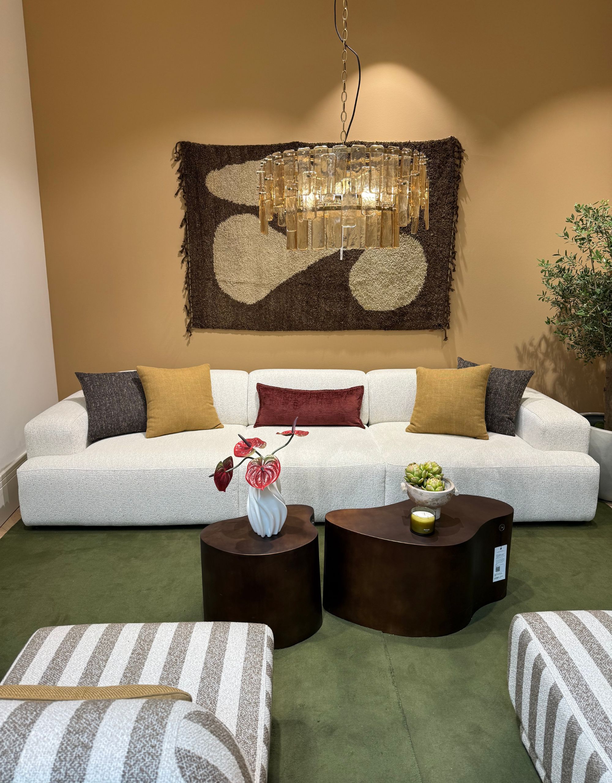 A living room setup in a showroom with a large white sofa with different colored scatter cushions in front of two organic shaped tables with a chandelier above and a rug as art on the wall behind