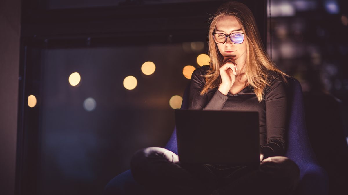 Woman sitting in a dark room with lights in the backgorund, using a computer which is lighting up her face