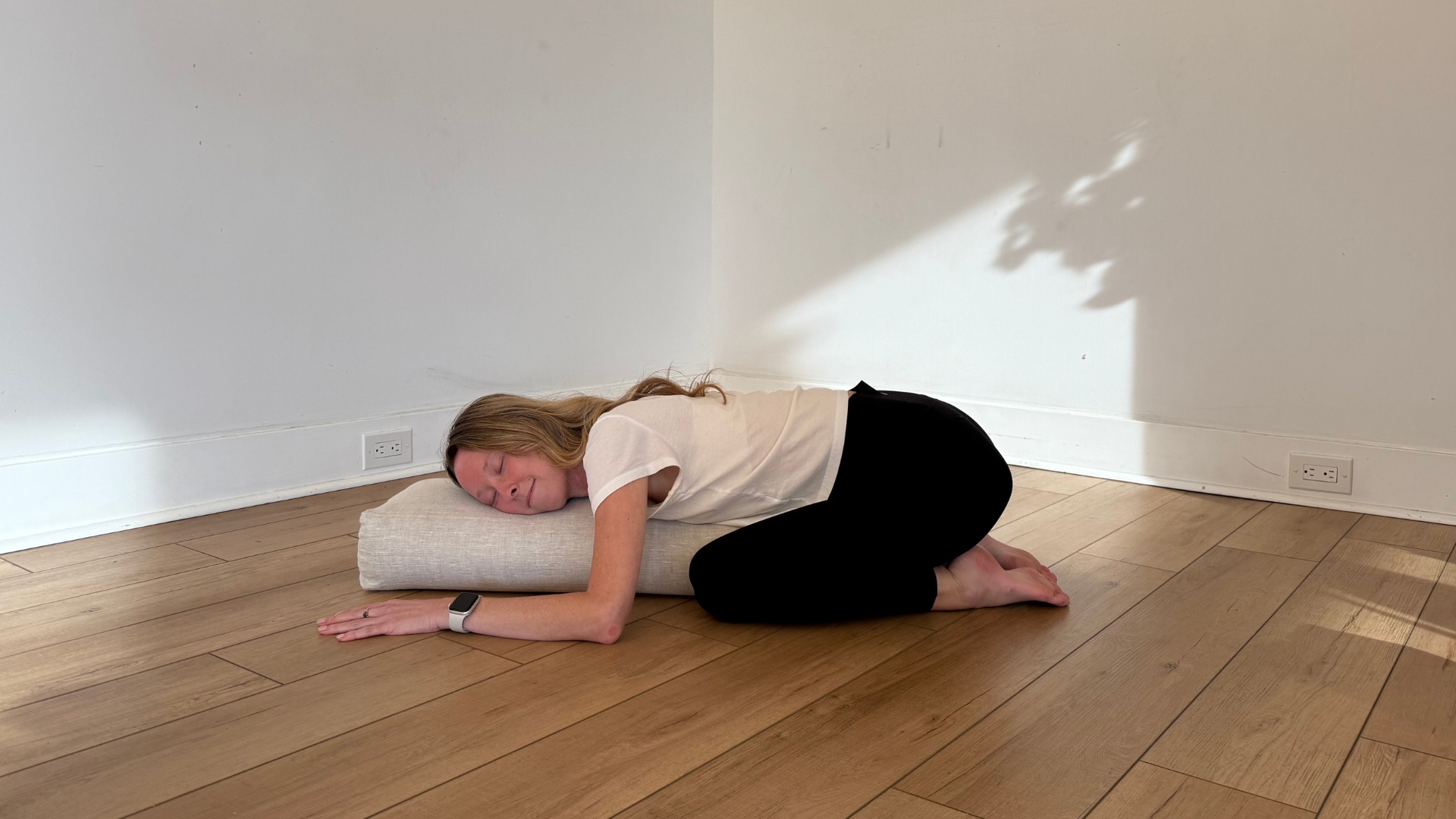 woman in child&#039;s pose folded over on a large cushion, on a wooden floor with white walls behind her