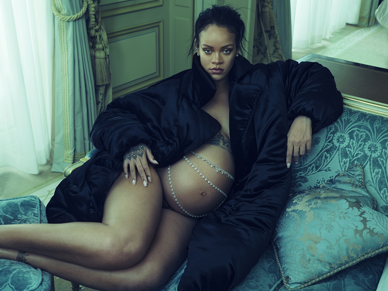 A pregnant Rihanna sits on a turquoise velvet chaise lounge, draped in a black fur coat with a silver chain draped across her bare stomach.