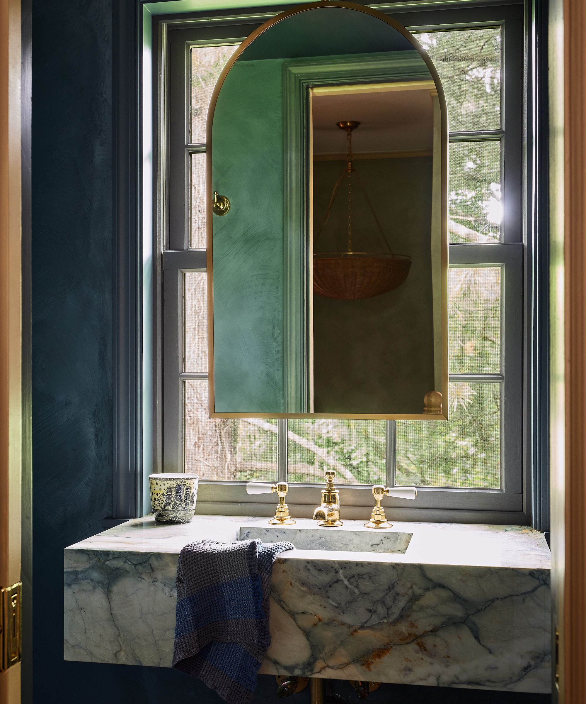 a blue limewashed bathroom with arched mirror hung in front of the window with a marble floating sink