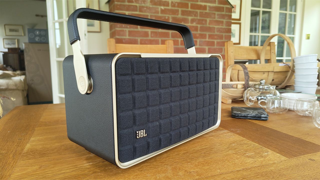 A black JBL Authentics 300 wireless speaker positioned on a wooden table. An exposed brick wall and two doors are visible in the background.