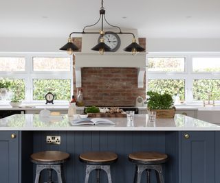 large kitchen with blue and white kitchen island, in front of exposed brick section of wall with white windows either side