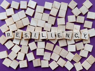 Square cubes with letters on them spell out the word "resiliency." These cubes sit on top of blank square cubes against a purple backdrop.