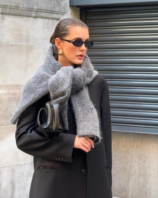 Fashion influencer Valeria @sobalera poses on a London sidewalk wearing a slicked-back bun, black oval sunglasses, chunky earrings, textured gray sweater tied over the shoulders, black blazer, and a black bag worn as a clutch.