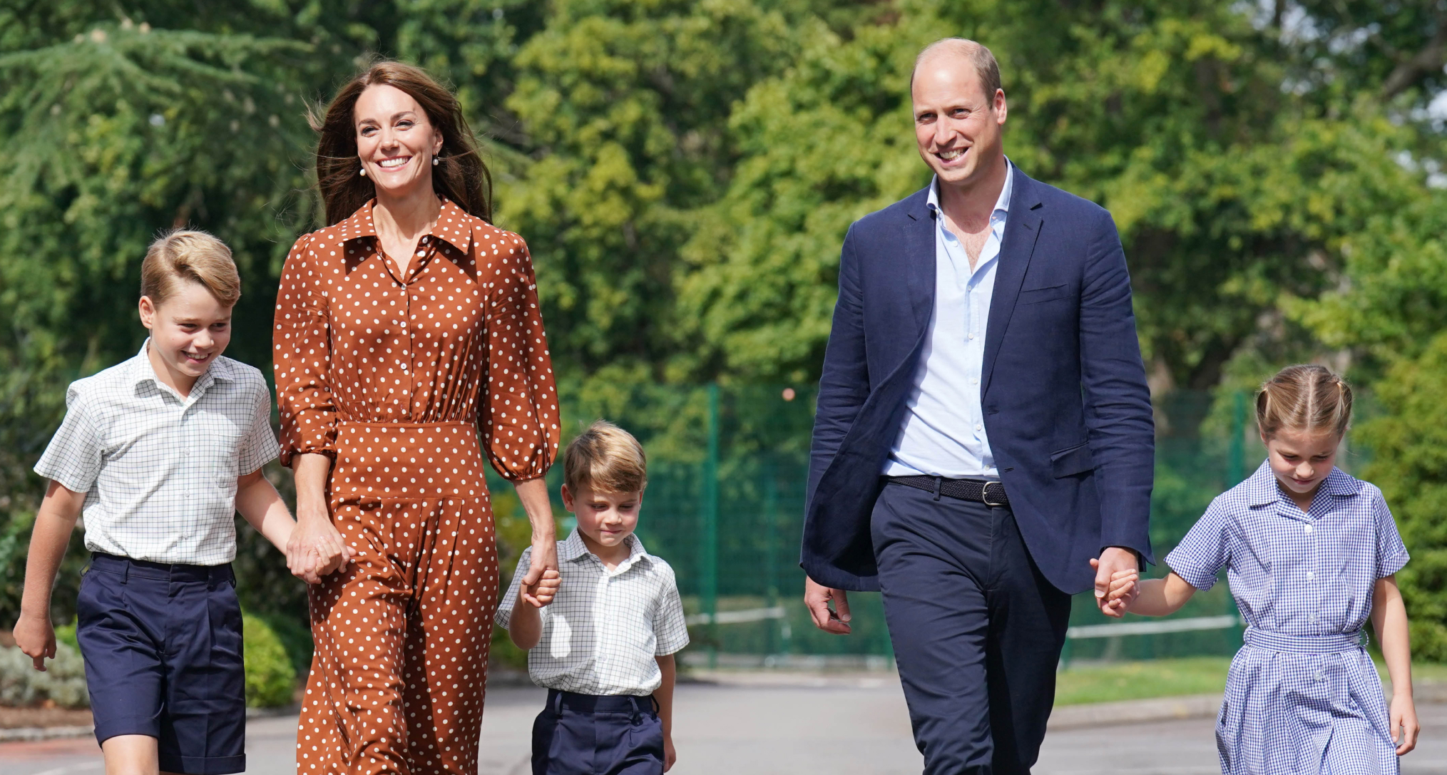 Princess Kate wearing a brown dress holding hands with Prince George and Prince Louis in school uniform walking next to Prince William holding hands with Princess Charlotte