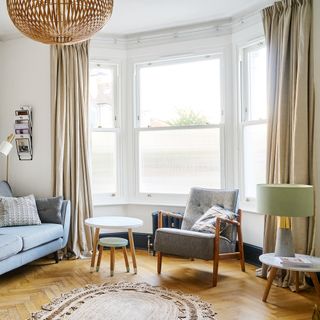 Living room with grey mid century style sofa and armchair, wooden herringbone parquet flooring, a nest of Scandi-style side tables and a matching table lamp