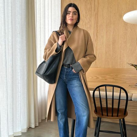 British fashion influencer Anna Howard poses in her chic wood-paneled London kitchen wearing a camel coat, gray sweater, black tote bag, black belt, and straight-leg jeans