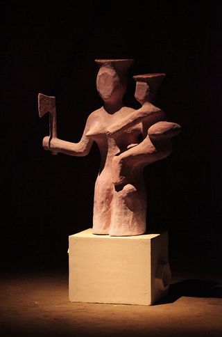 A sculpture of a woman with a child in one hand and an ax in the other on a square platform.