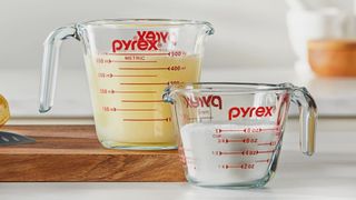 Two Pyrex measing jugs in a kitchen - one is filled with yellow liquid and measures in mtric, the other is filled with white liquid and measures in cups