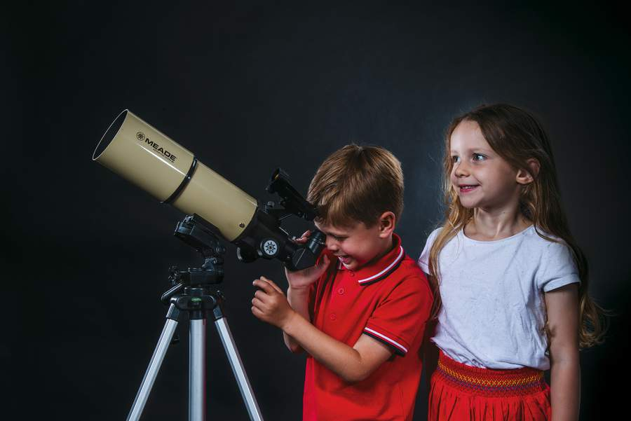 Two children using a telescope. One is looking through the telescope while another looks up.