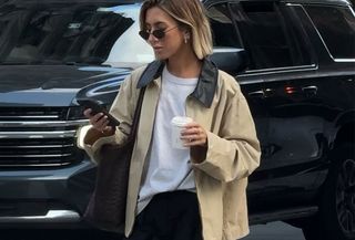 Woman in barn jacket, t-shirt, and black pants looking at a phone.