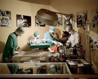 View of a diorama at Chris Barnard Museum - the scene is of an operating room with hospital staff dressed in white and green and wearing hair protectors, masks and gloves. There are multiple black and white images on the beige coloured walls and there is a man tending to the diorama