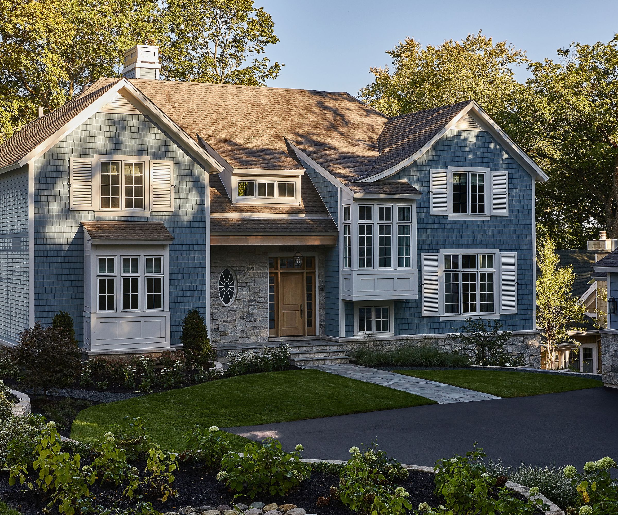 A home exterior with muted blue brickwork, white trim and window shutters, and a wooden front door. A front garden with lawn and plants, and a paved path.