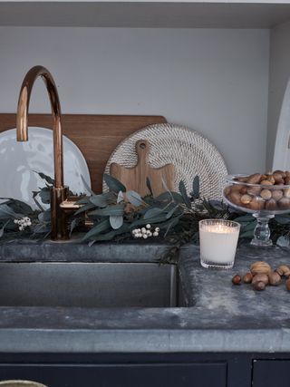 detail image of a zinc countertop and sink with copper faucet, bowl with nuts, candle, chopping boards, eucalyptus and berries behind the faucet