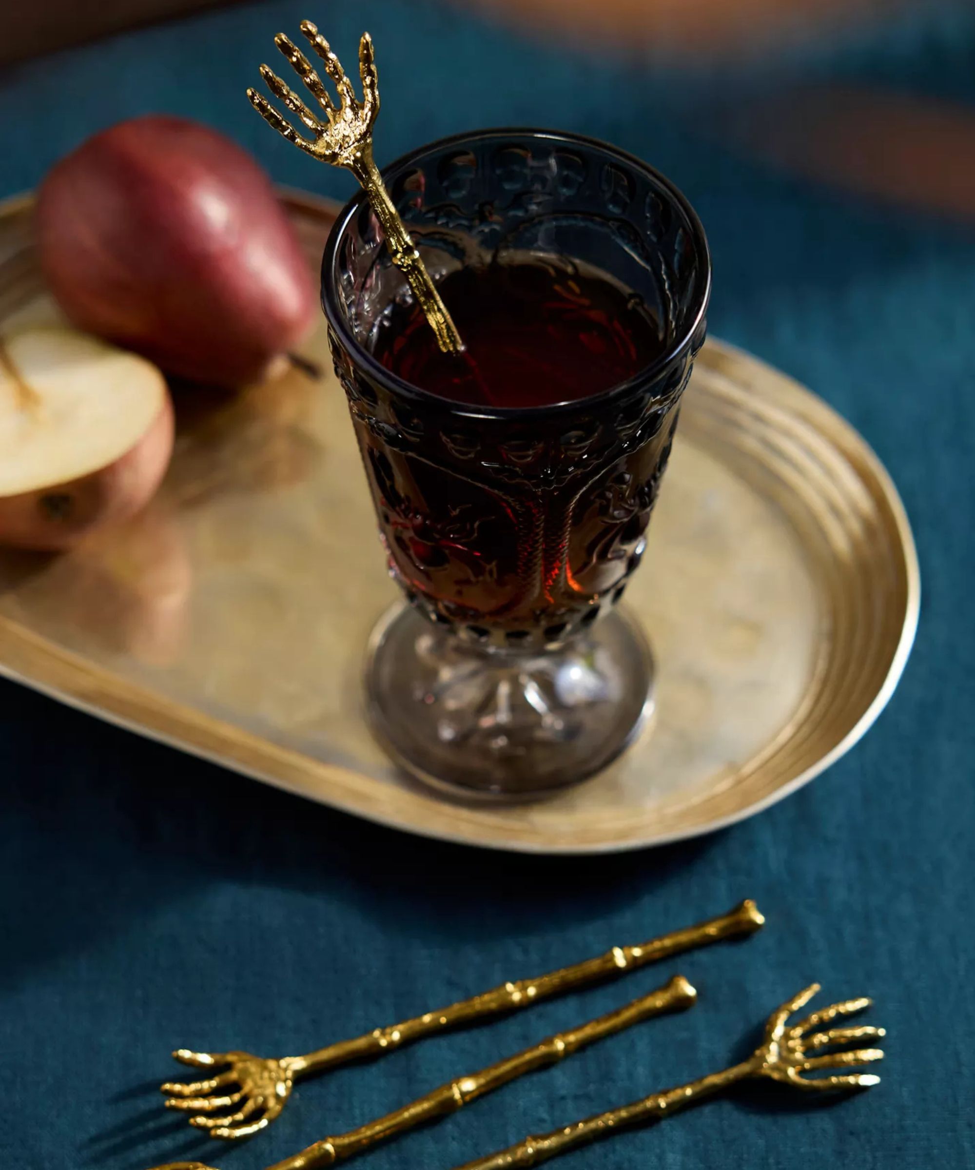 Teal blue tablecloth with three brass skeleton-shaped drinks stirrers, a brass tray and a grey drinking glass