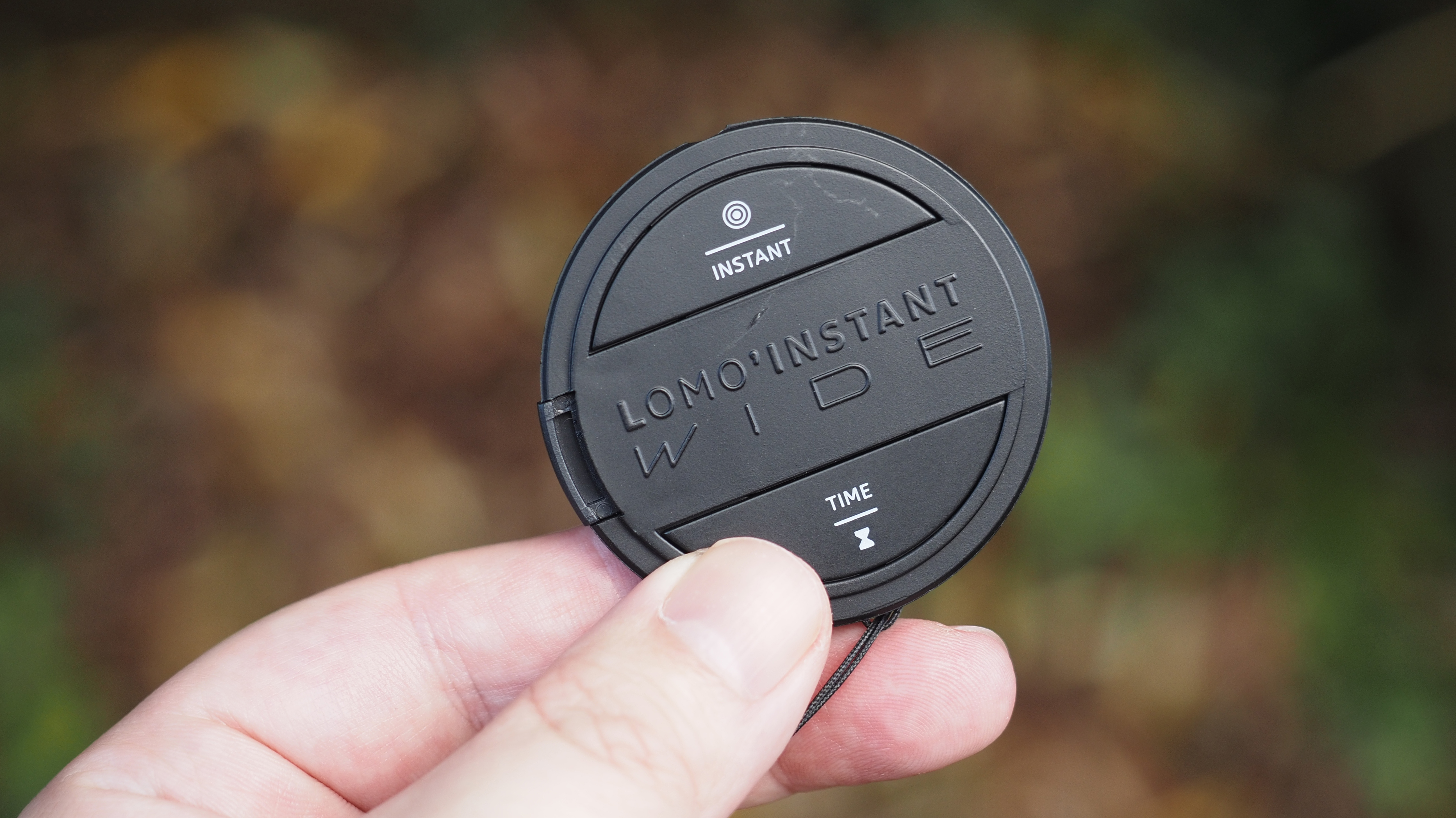 A hand holding the lens cap/remote control of the Lomo'Instant wide glass camera, against a forest background