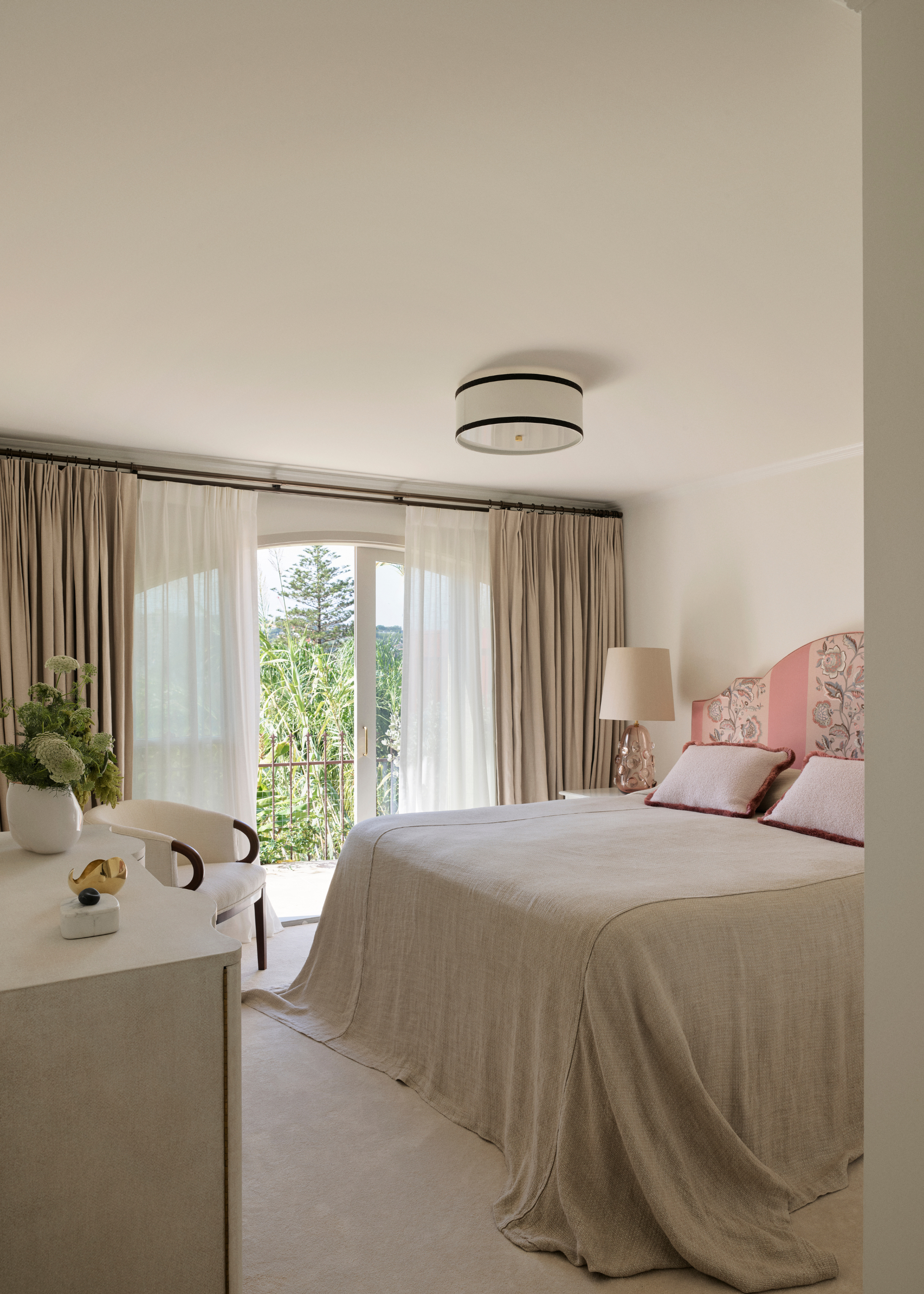 An off-white minimalist bedroom with muted window treatments, a sideboard, a sofa chair, a patterned headboard and a ceiling light