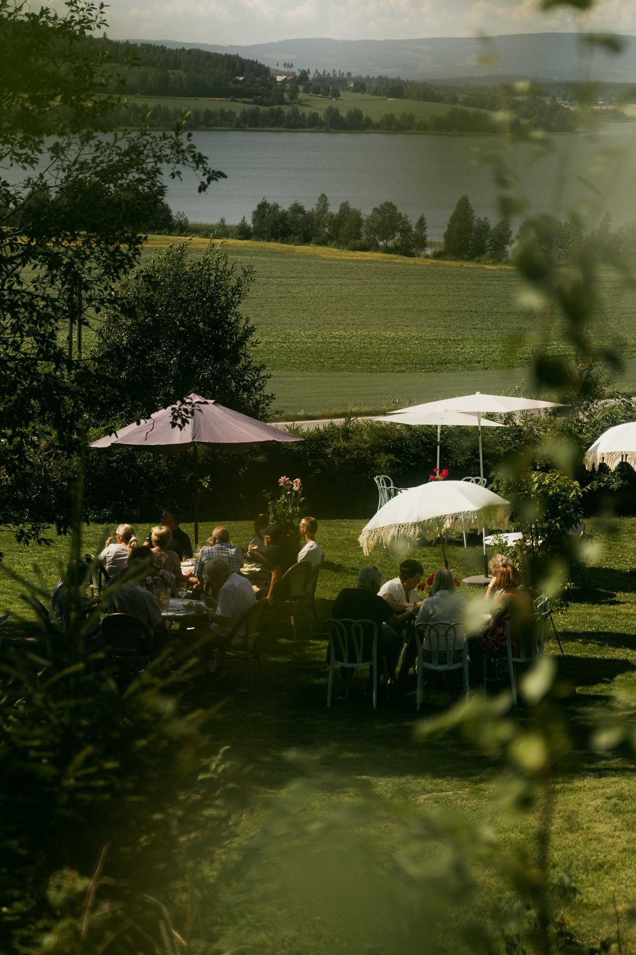 A bucolic landscape, showing verdant vegetation and a lake, is decorated with retro-inspired parasols as a crowd of people enjoys their time outdoors.