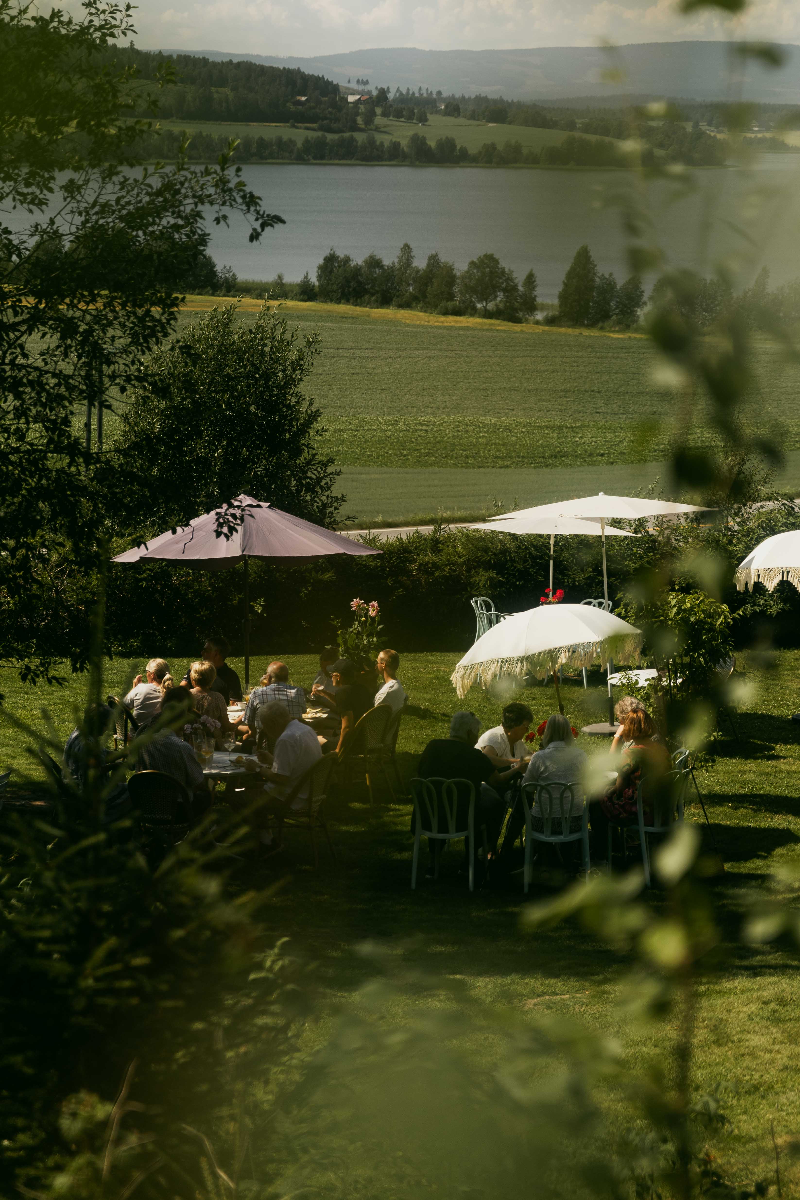 A bucolic landscape, showing verdant vegetation and a lake, is decorated with retro-inspired parasols as a crowd of people enjoys their time outdoors. 