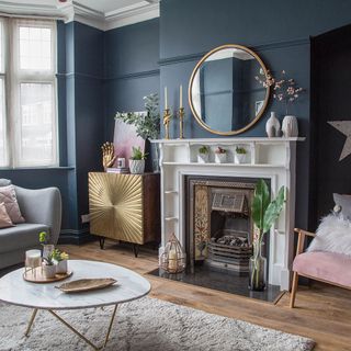 a living room with dark blue walls, fireplace with white mantel, gold patterned unit, wooden floors with a rug and gold marble coffee table on top