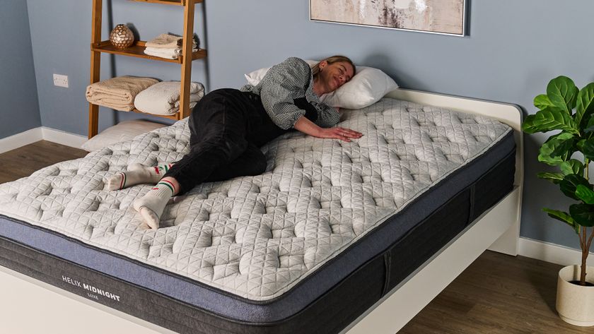 The image shows a woman wearing black dungarees lying on her side on the Helix Midnight Luxe mattress, which is placed on a white bedframe in a blue bedroom