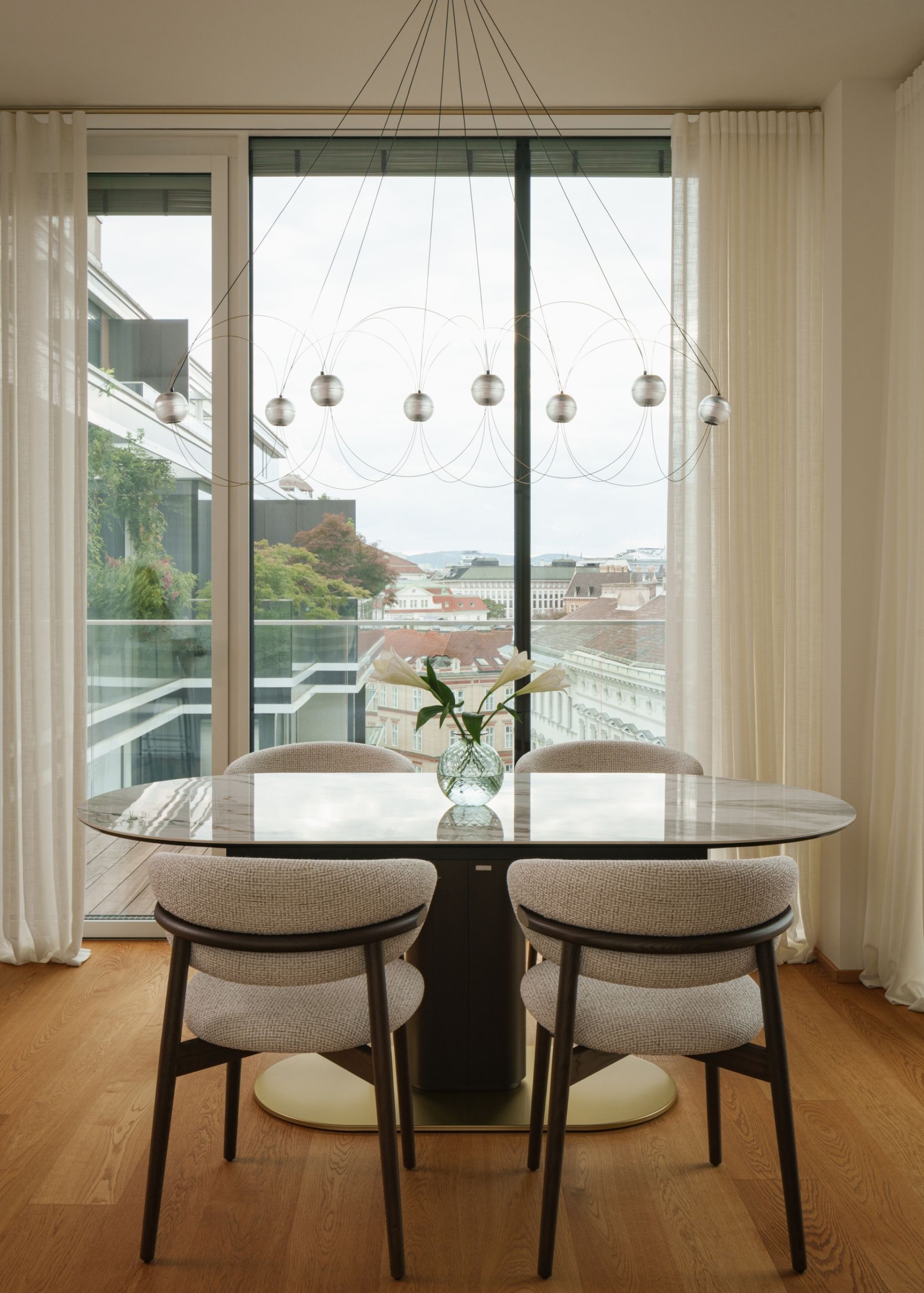 Image of a dining area with wood floors, white curtains, white walls, and large windows showing a city outside. There is an oval marble table with white boucle chairs around it. 