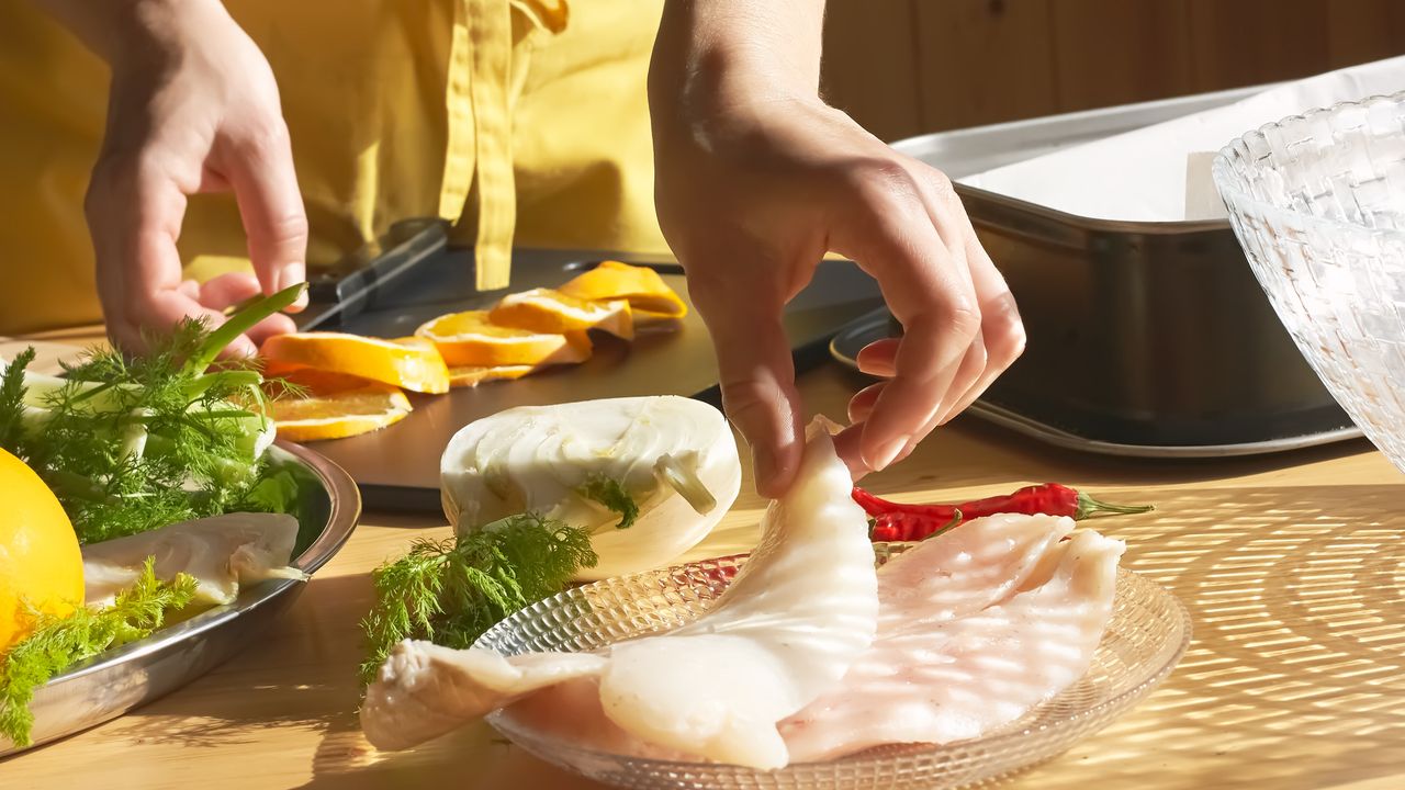 Hands pick up white fish filet from glass plate. Various herbs, vegetables cooking utensils surround the fish