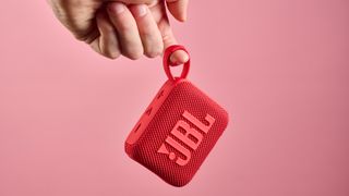 A red JBL Go 4 is being held in the air from the corner loop in front of a pink background.