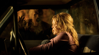 A woman sits in a car and looks scared while a ghost peers through the window