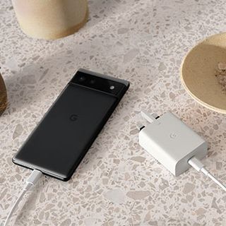 A Google Pixel 6 face-down on a surface, connected to a wall charger with an engraved Google "G" logo